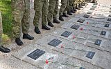 Uczczenie pamięci  ofiar wojny na cmentarzu, po lewej stronie widać nogi stojących żołnierzy, na płytach leżących na ziemi ciemne płyty z nazwiskami poległych i datami, na płytach po jednym czerwonym goździku