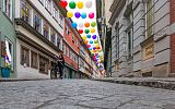 uliczka z zawieszonymi między kamienicami kolorowymi balonami. kobieta prowadzi rower.