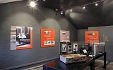 wystawa w centrum imigracyjnym: na tle ciemnoszarych ścian pomarańczowe plansze ze zdjęciami, na środku gablota i otwarta walizka z eksponatami