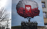 Ściana szkoły a na niej mural. Malowidło przedstawia Powstańców którzy stoją pod drzewem. Korona drzewa w barwach biało czerwonych - symbolizuje flagę Polski.