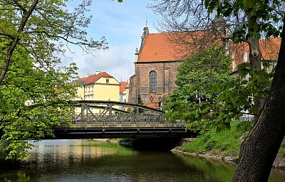 Most z ozdobnymi łukami nad rzeką, po bokach drzewa, w tle kościół z czerwonym dachem